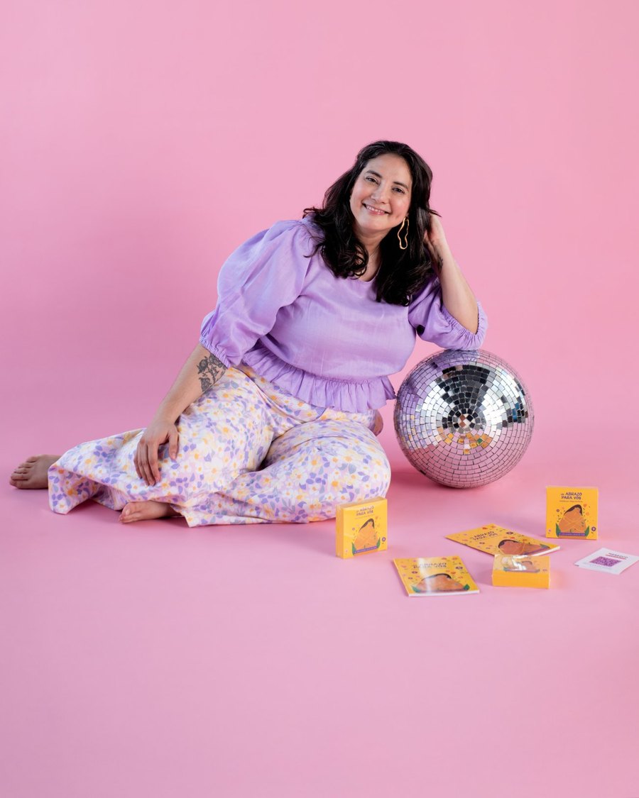 Ale sitting on a pink floor next to a disco ball and her Un abrazo para vos affirmation cards